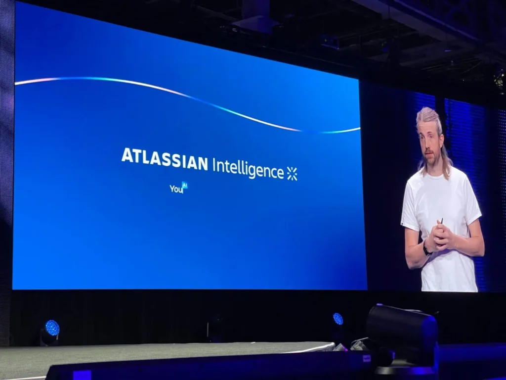 A presentation on atlassian intelligence witho a keynote speaker next to a large screen.